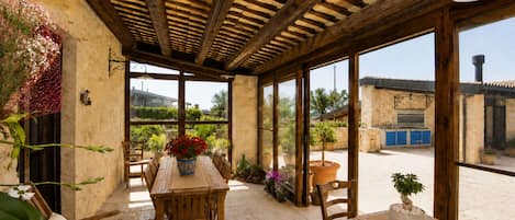 A covered patio with a wooden table and chairs, surrounded by potted plants and a view of the countryside.