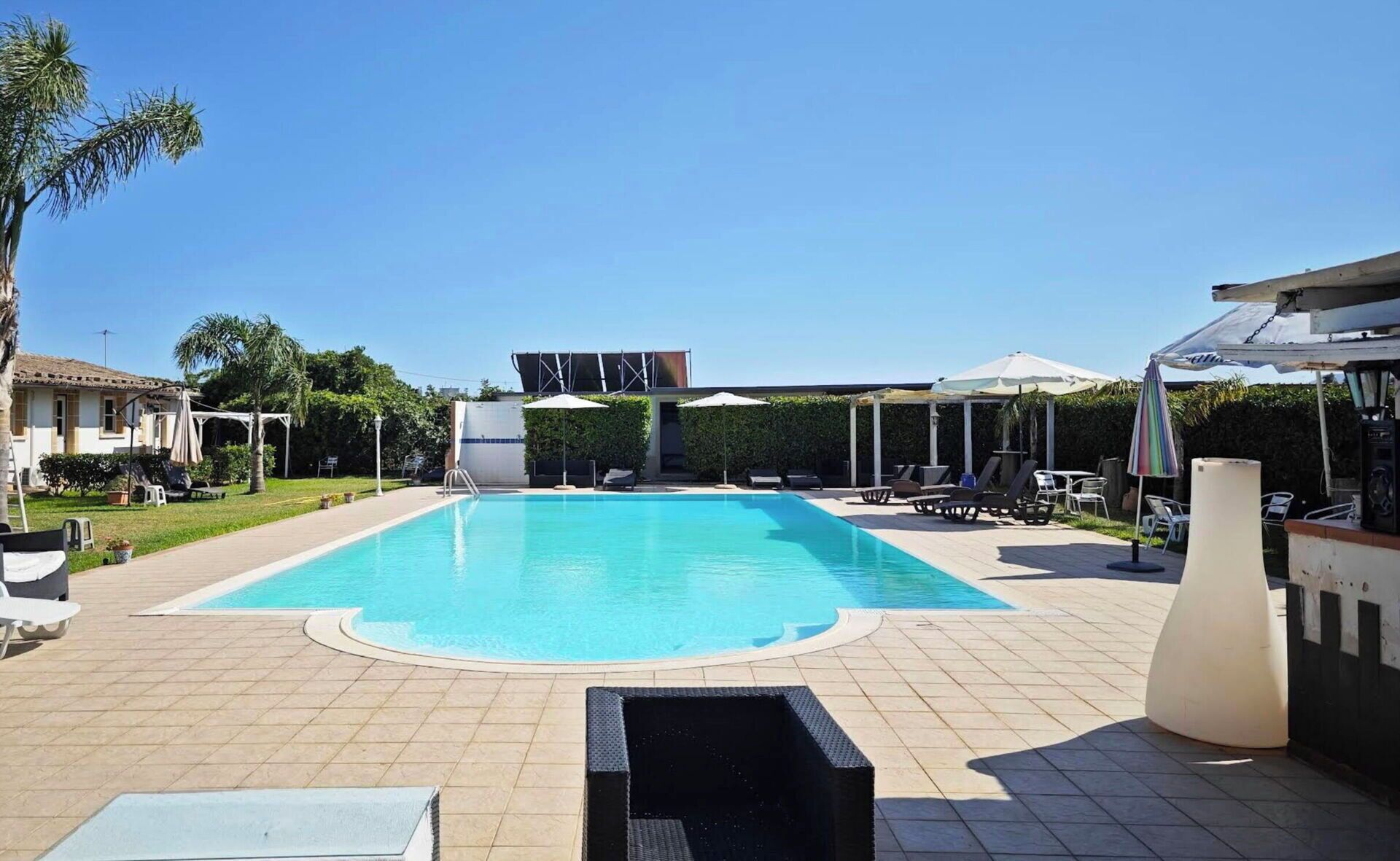 Piscina com espreguiçadeiras e guarda-sóis, rodeada por um deck revestido e vegetação.