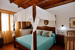 Ein Zimmer mit einem Himmelbett, Holzbalkendecke, einem kleinen Schrank und einem Fenster mit Vorhängen.