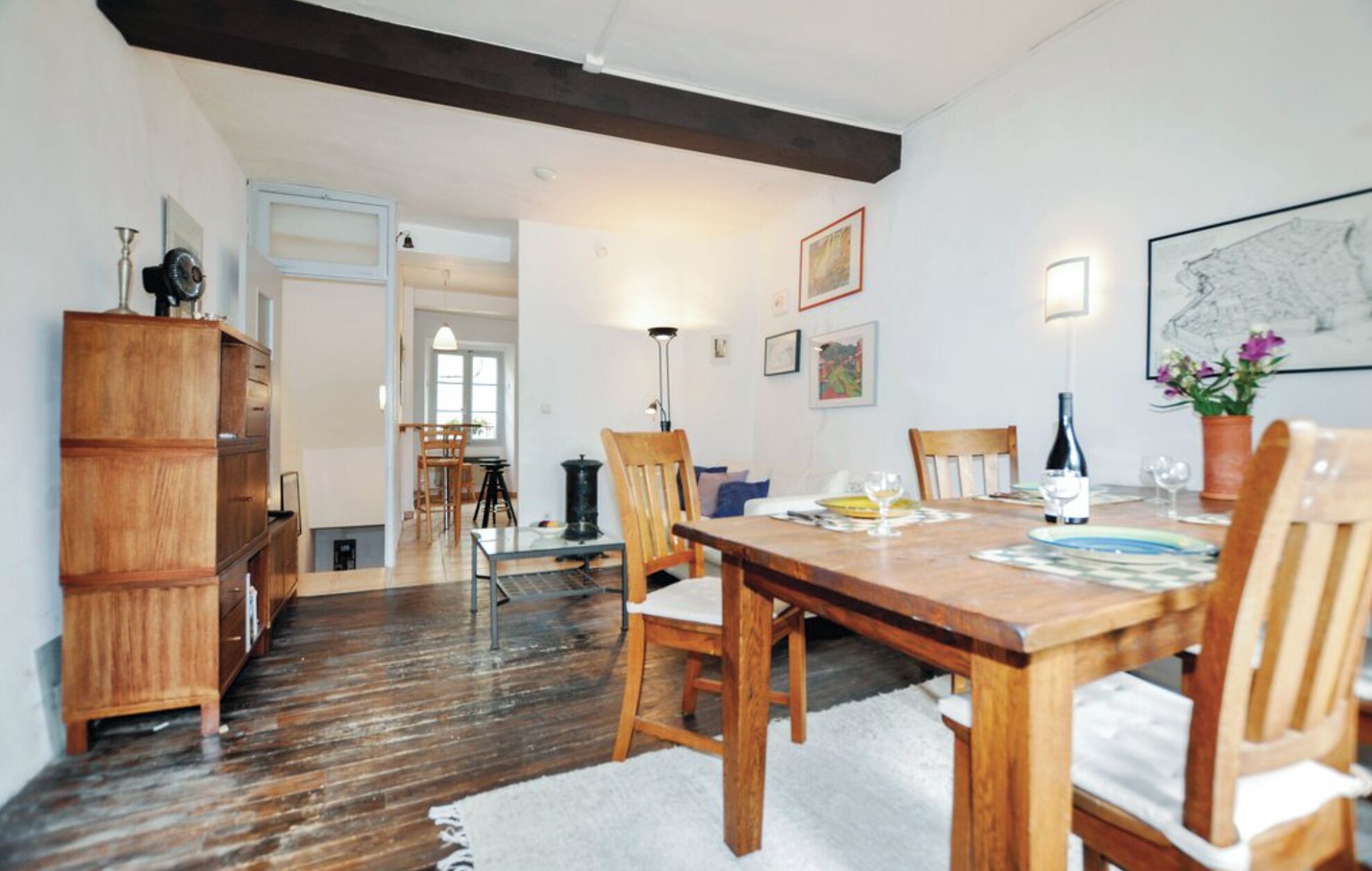 Un coin repas avec une table en bois, des chaises et un tapis blanc. Un buffet en bois se trouve à gauche, et une porte donne accès à une autre pièce.