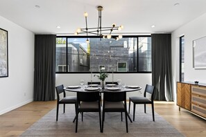 Comedor moderno con una mesa redonda preparada para cuatro personas, una araña y ventanas amplias con cortinas.