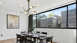 A dining area with a round table, black chairs, a chandelier, and a large window with a view of buildings. - Rooftop + Grill | Venice Canals + Abbot Kinney | Canal Crown (Los Angeles)