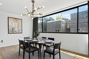 A dining area with a round table, black chairs, a chandelier, and a large window with a view of buildings. - Rooftop + Grill | Venice Canals + Abbot Kinney | Canal Crown (Los Angeles)
