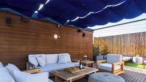 An outdoor seating area with a wooden deck, a covered section with blue fabric, and a wooden wall with a clock. - Rooftop + Grill | Venice Canals + Abbot Kinney | Canal Crown (Los Angeles)