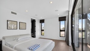 A modern bedroom with a large bed, two framed artworks, a window with curtains, and a door leading to another room. - Rooftop + Grill | Venice Canals + Abbot Kinney | Canal Crown (Los Angeles)