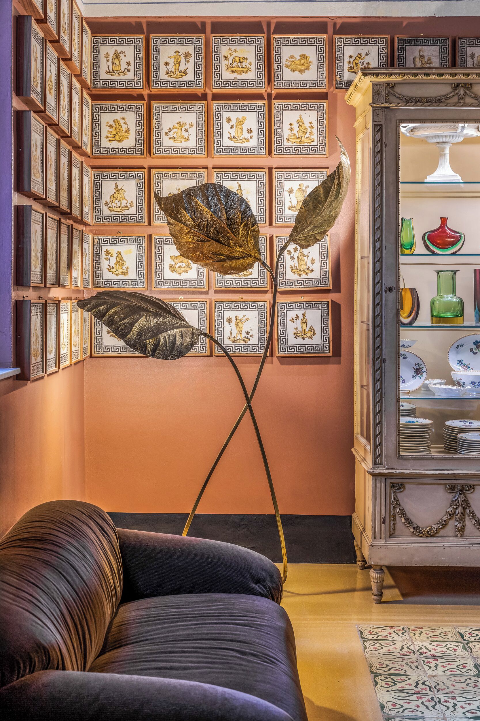 Una habitación con sofá, una lámpara de pie decorativa, una pared con obras de arte enmarcadas y un armario con vajilla.