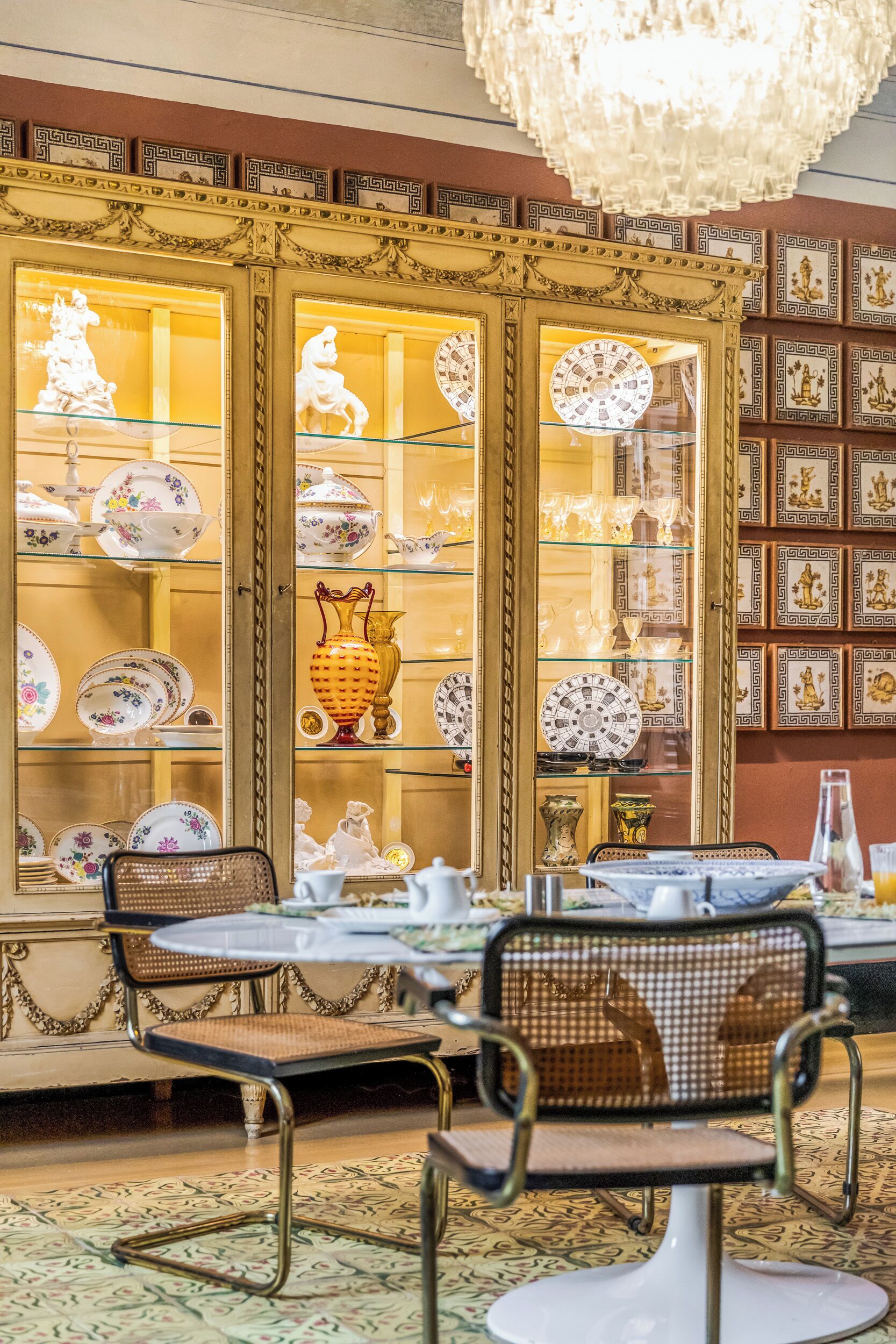 A dining area with a table set for two, surrounded by ornate chairs, and a display of fine china and glassware behind glass doors.
