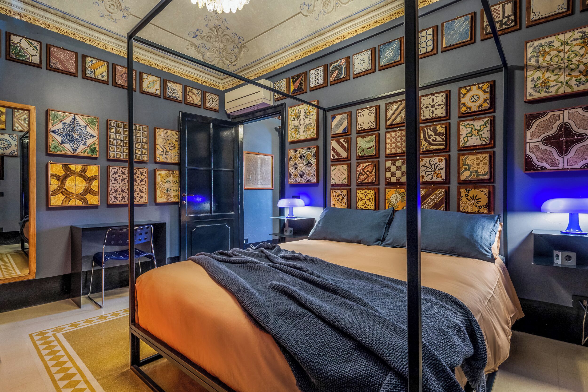 Una habitación con cama con dosel, un escritorio y una silla. Las paredes están decoradas con azulejos enmarcados.