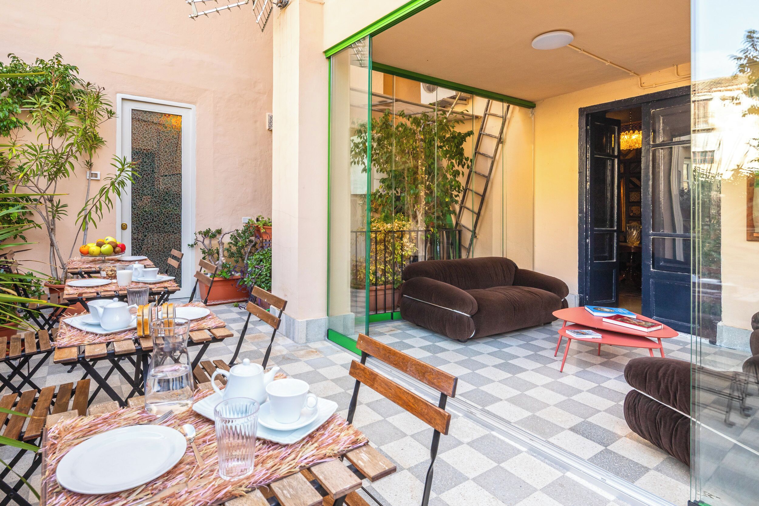 Ein gemütlicher Außenbereich mit Tischen, Stühlen und einer Couch, umgeben von Topfpflanzen und einer Glastür zum Balkon.