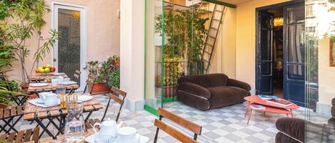 A cozy outdoor seating area with tables, chairs, and a sofa, surrounded by potted plants and a glass door leading to a balcony.