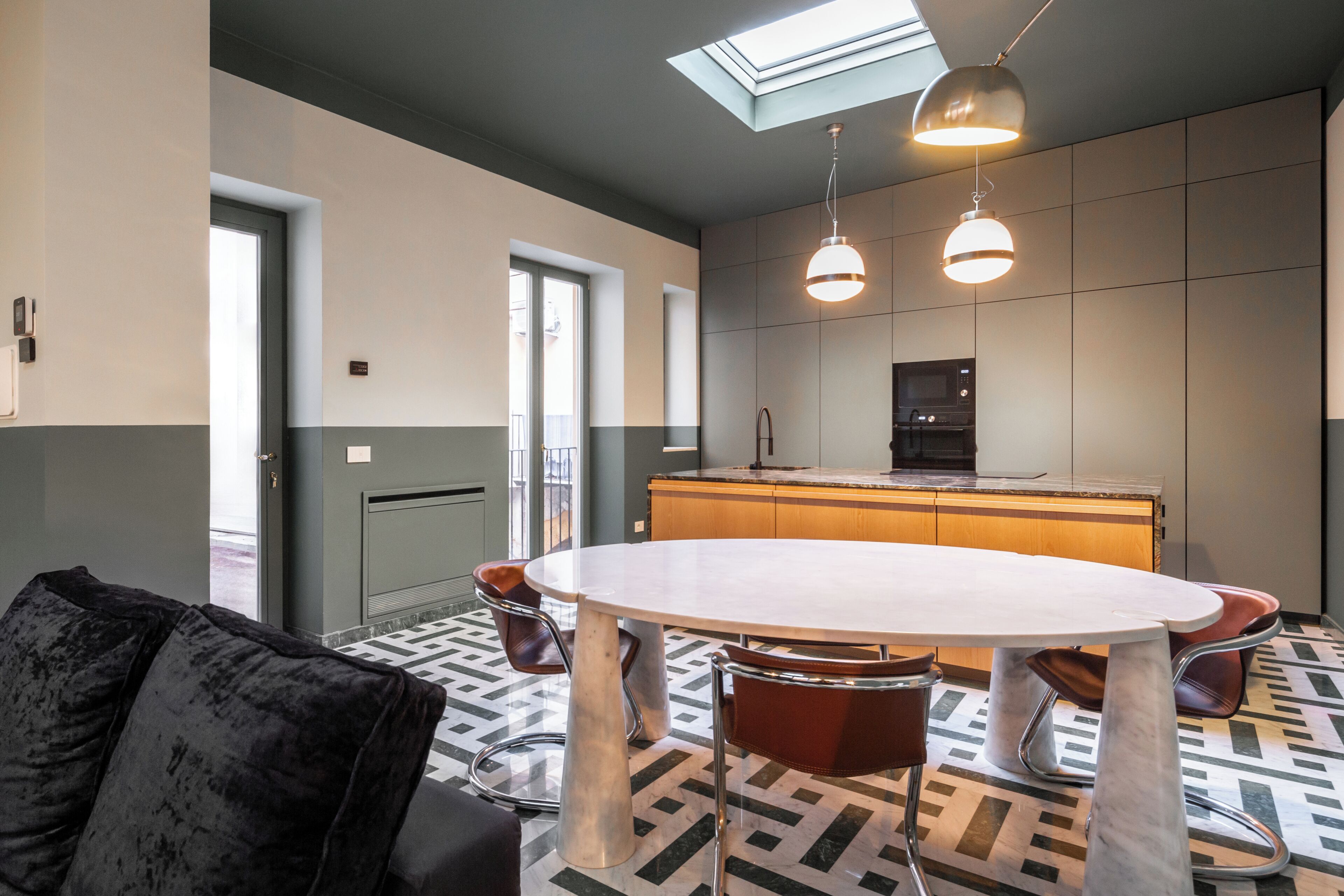 A modern kitchen with a microwave, a coffee machine, and a sink. There is a round dining table with chairs and a black sofa. The floor has a patterned tile design.