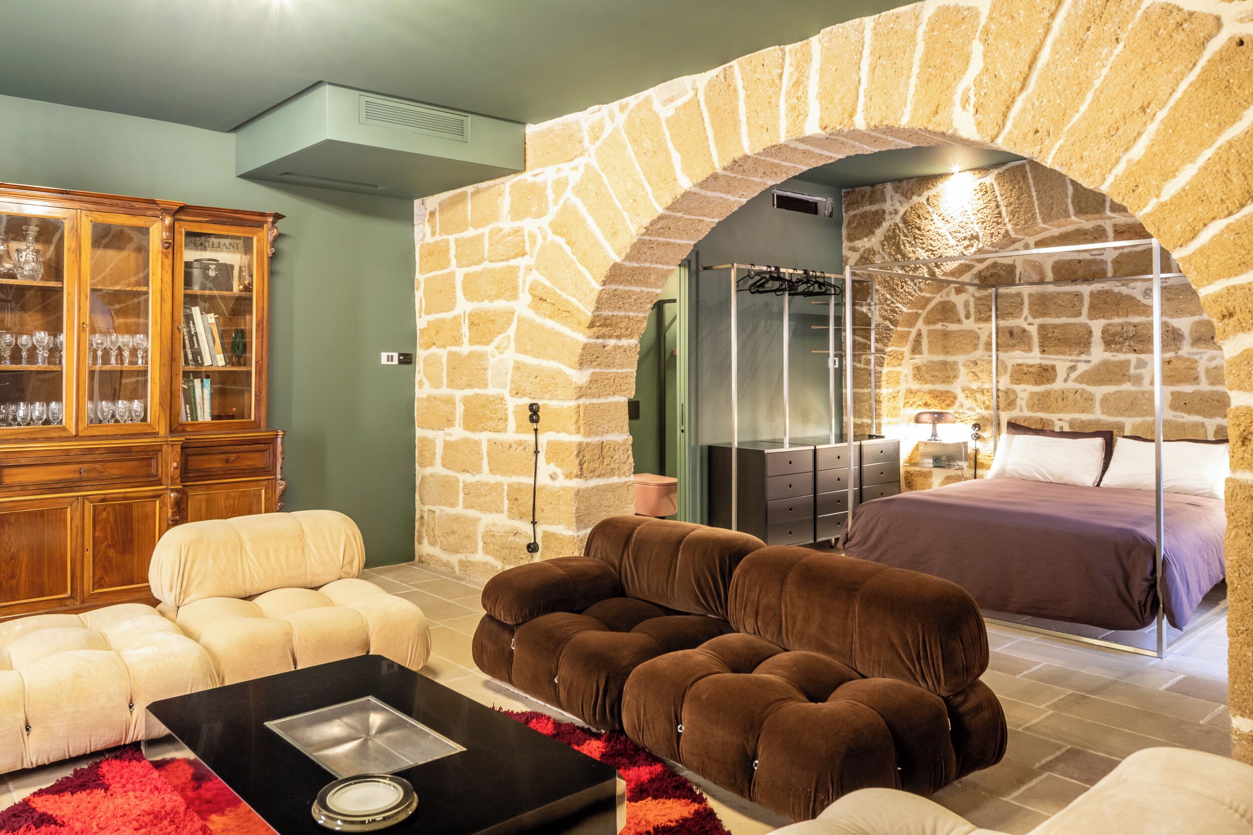 A cozy room with a stone archway, a bed, a sofa, a cabinet with glass doors, and a fireplace.