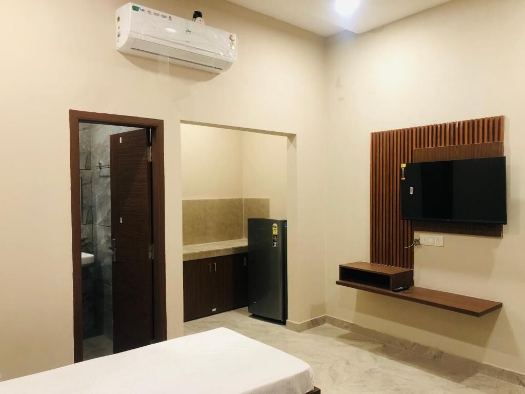 A compact hotel room with a bed, a small kitchenette, a built-in TV, and an air conditioning unit.