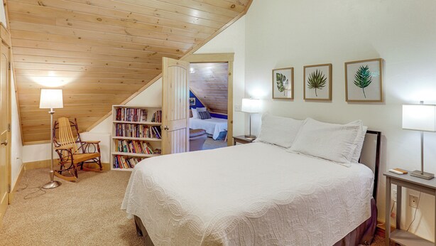 Un dormitorio con techo de madera, cama con ropa de cama blanca, una estantería y grabados botánicos enmarcados en la pared.