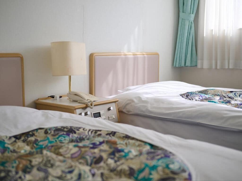 Habitación de hotel con dos camas, una mesita de noche con lámpara y una ventana con cortinas.