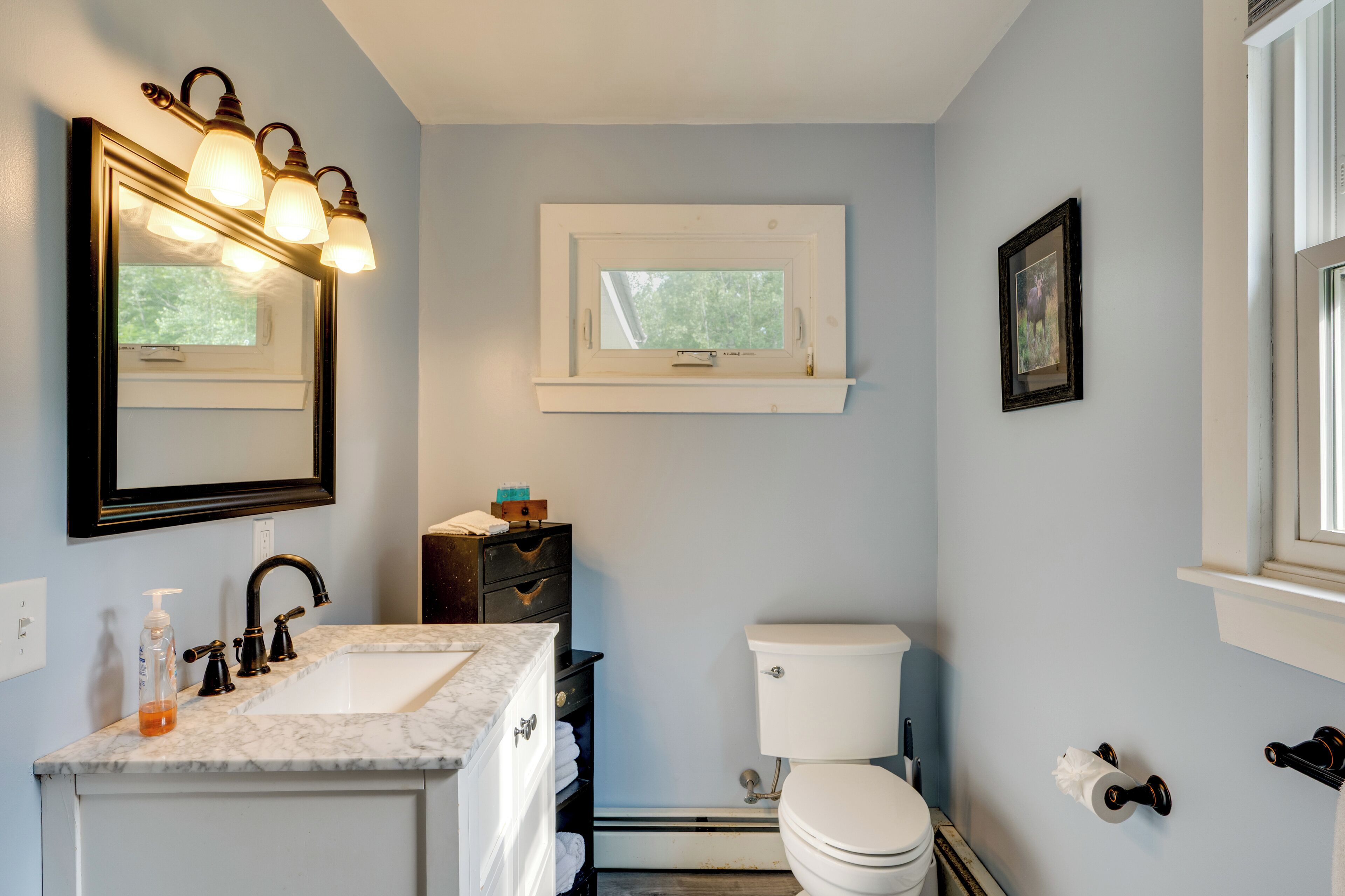 A bathroom with a sink, mirror, toilet, and window.