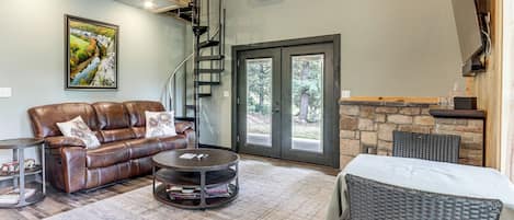 A spacious living area with a leather sofa, a round coffee table, and a wicker chair. There is a staircase leading to an upper level, and a large window with a view of trees.
