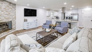 Ein modernes Wohnzimmer mit Kamin, Flachbildfernseher, Essbereich und einer Küche im Hintergrund sowie eine Sitzecke mit gemusterten Sofas.