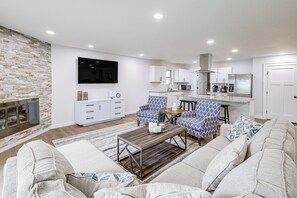 A modern living room with a fireplace, a flat-screen TV, a dining area with a kitchen in the background, and a seating area with patterned sofas. - Lake-View Deck! Modern Greers Ferry Lake Retreat (Higden)