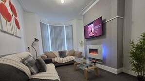 A modern living room with a sofa, a glass coffee table, a flat-screen TV mounted on the wall, and a small plant. - Cozy House in Liverpool (Liverpool)