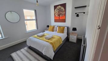 A bedroom with a bed, a bedside table, a lamp, a radiator, a window, and a wall-mounted artwork.