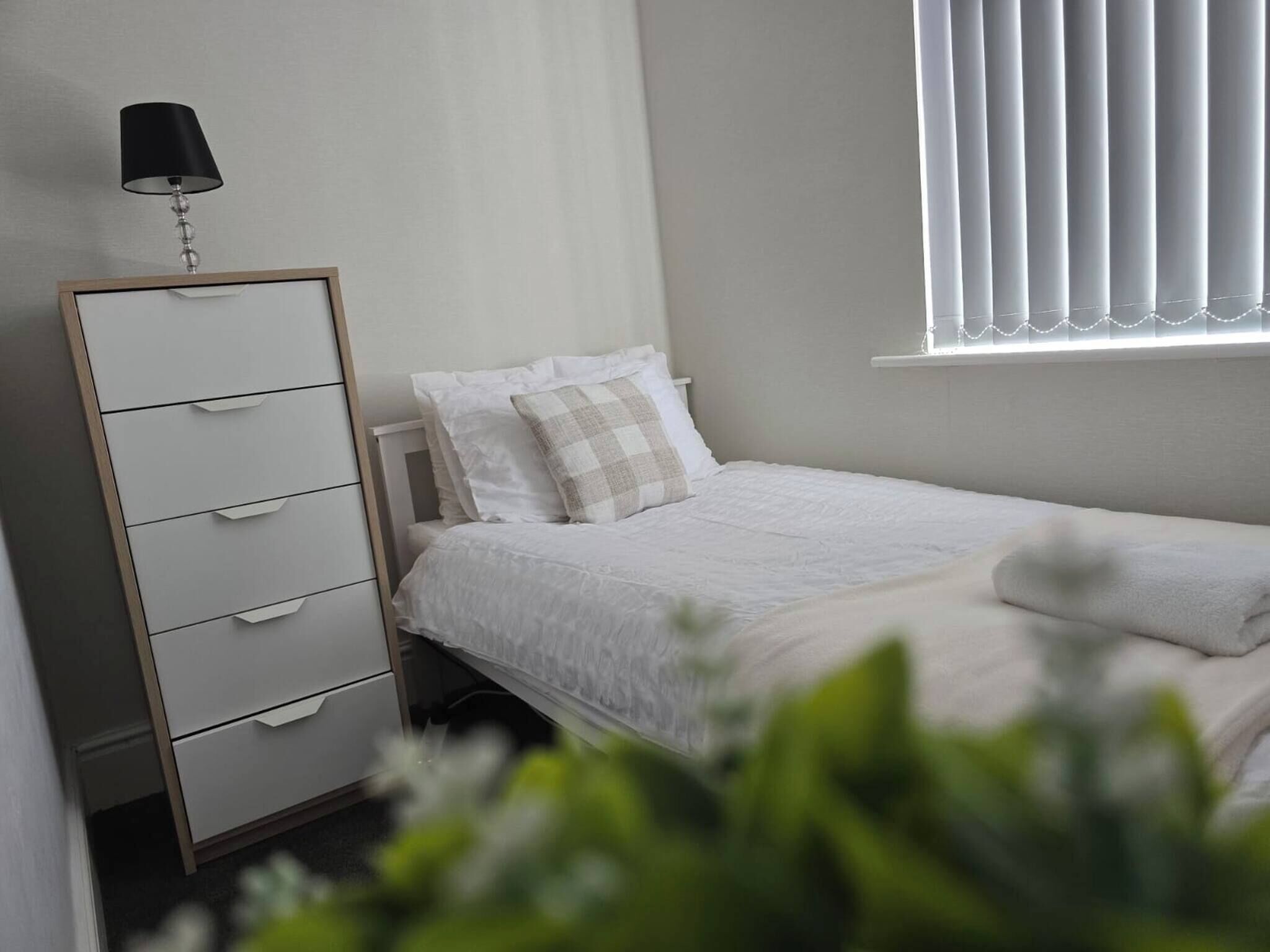 A bedroom with a bed, a dresser, a lamp, and a window with blinds.