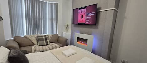 A modern hotel room with a bed, a sofa, a flat-screen TV, and a fireplace.