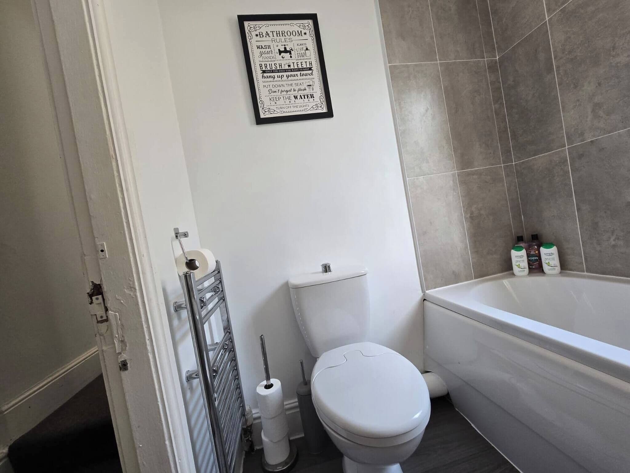 A bathroom with a toilet, a roll of toilet paper, a metal rack, and a framed sign on the wall.
