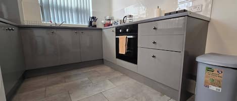 A kitchen with grey cabinets, a black oven, a window with blinds, and a grey trash bin.
