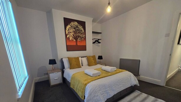 A bedroom with a bed, nightstands, a radiator, a wall-mounted coat rack, and a painting of a tree. - Cozy House in Liverpool (Liverpool)