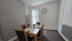 A dining room with a wooden table set for four, a clock on the wall, and a window with blinds. - Cozy House in Liverpool (Liverpool)