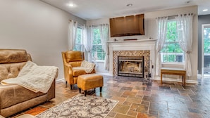 A living room with a fireplace, leather sofa, ottoman, patterned rug, and a large flat-screen TV. - Treehouse Views! Walking Distance to IMS (Indianapolis)