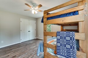 Un lit superposé avec une couverture à motifs, un ventilateur de plafond en bois et une porte blanche dans une pièce dont le sol est recouvert de planches de bois.