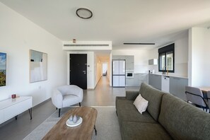 A modern living room with a sofa, armchair, and coffee table, leading to a kitchen and dining area. - Stylish 4BR Apt l Balcony & Mamad (Bat Yam)