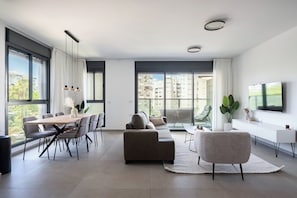A modern living room with a dining area, large windows, a flat-screen TV, and a view of buildings. - Stylish 4BR Apt l Balcony & Mamad (Bat Yam)