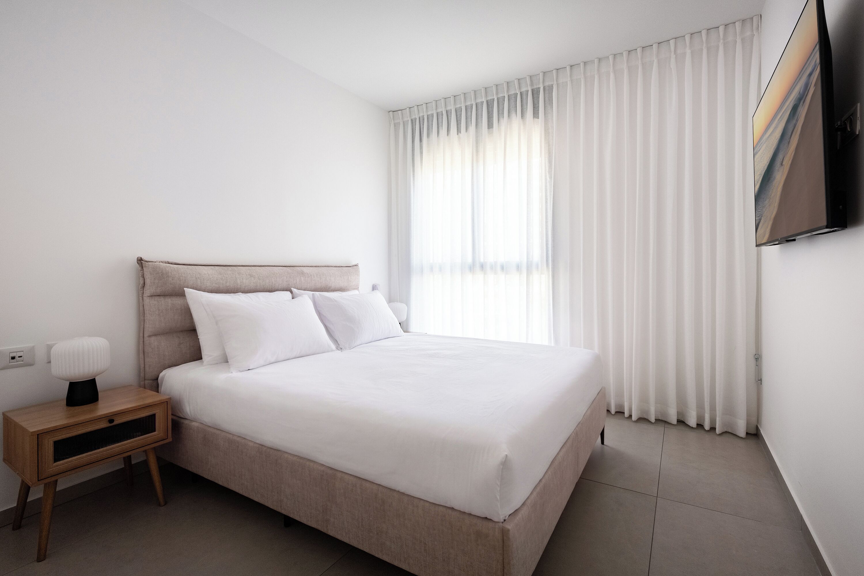 A modern hotel room with a bed, a nightstand, a flat-screen TV, and a large window with curtains.