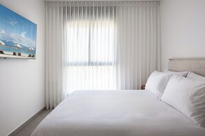 Hotellihuone, jossa on suuri sänky, valkoiset verhot ja taulu, jossa on kuvattuna rantamaisema.