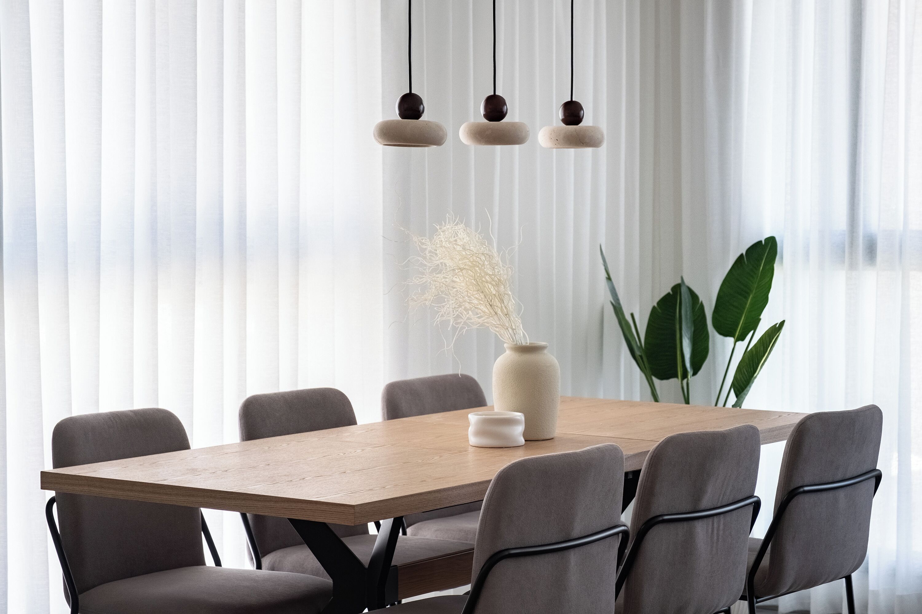 Un coin repas avec une table en bois, six chaises, un vase contenant des plantes séchées et des lampes suspendues.