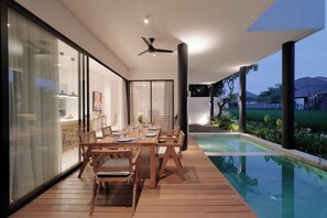 A modern outdoor dining area with a pool, wooden deck, and glass walls. - VOX Residences Canggu (Bali)