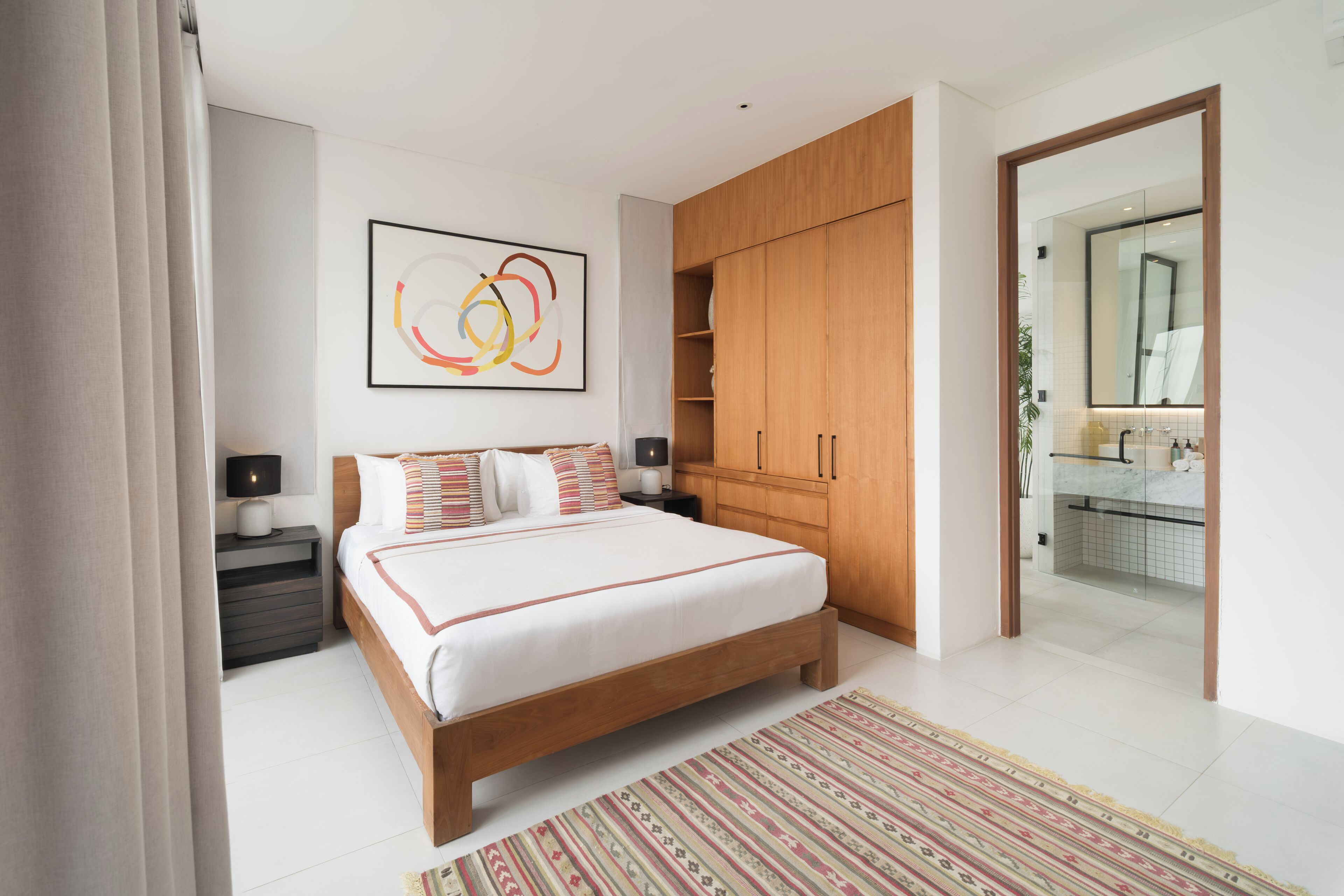 A modern bedroom with a large bed, wooden wardrobe, bedside tables, and a bathroom visible through an open door.