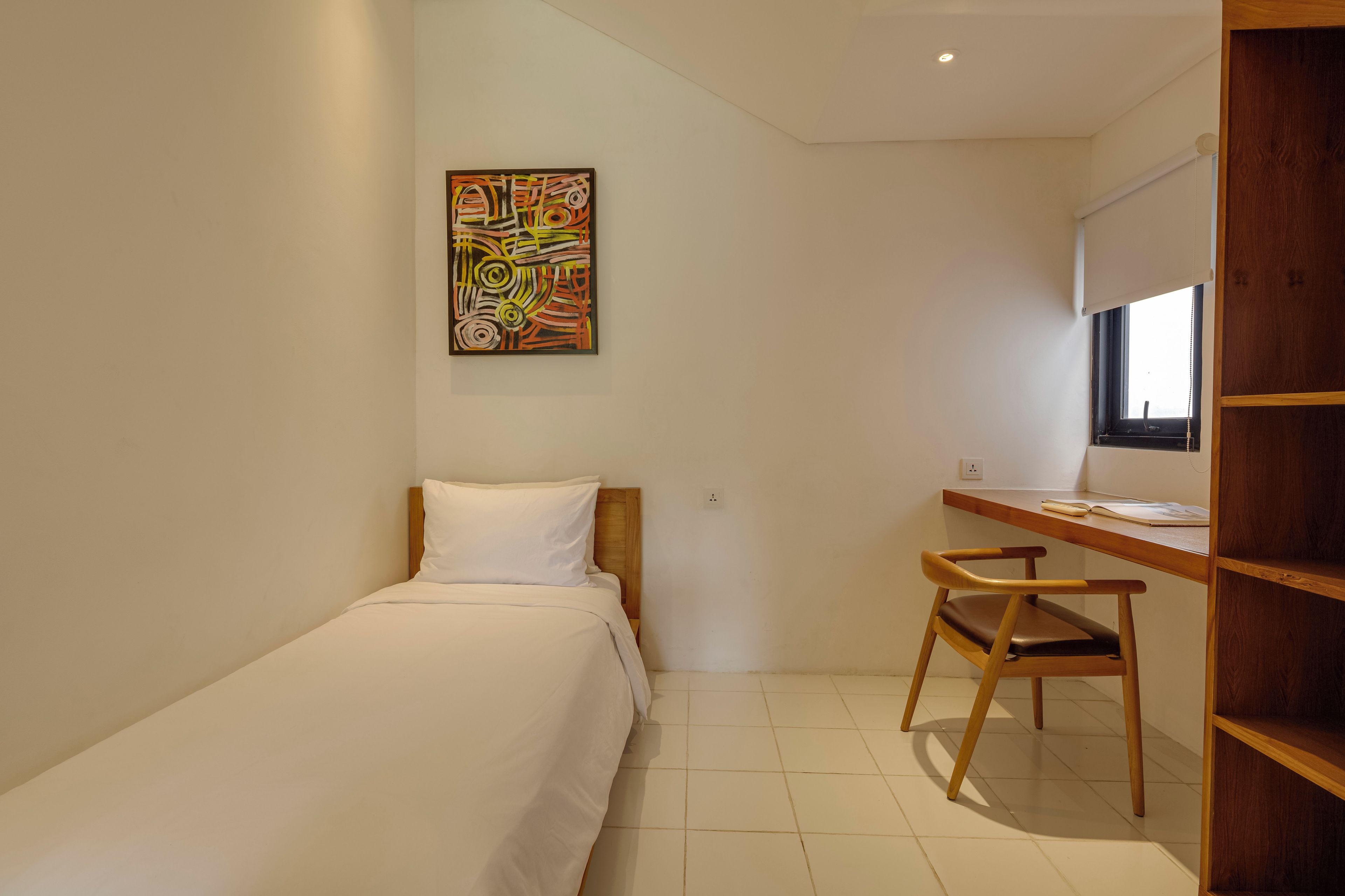 A small, neatly arranged bedroom with a single bed, a desk with a chair, and a window.