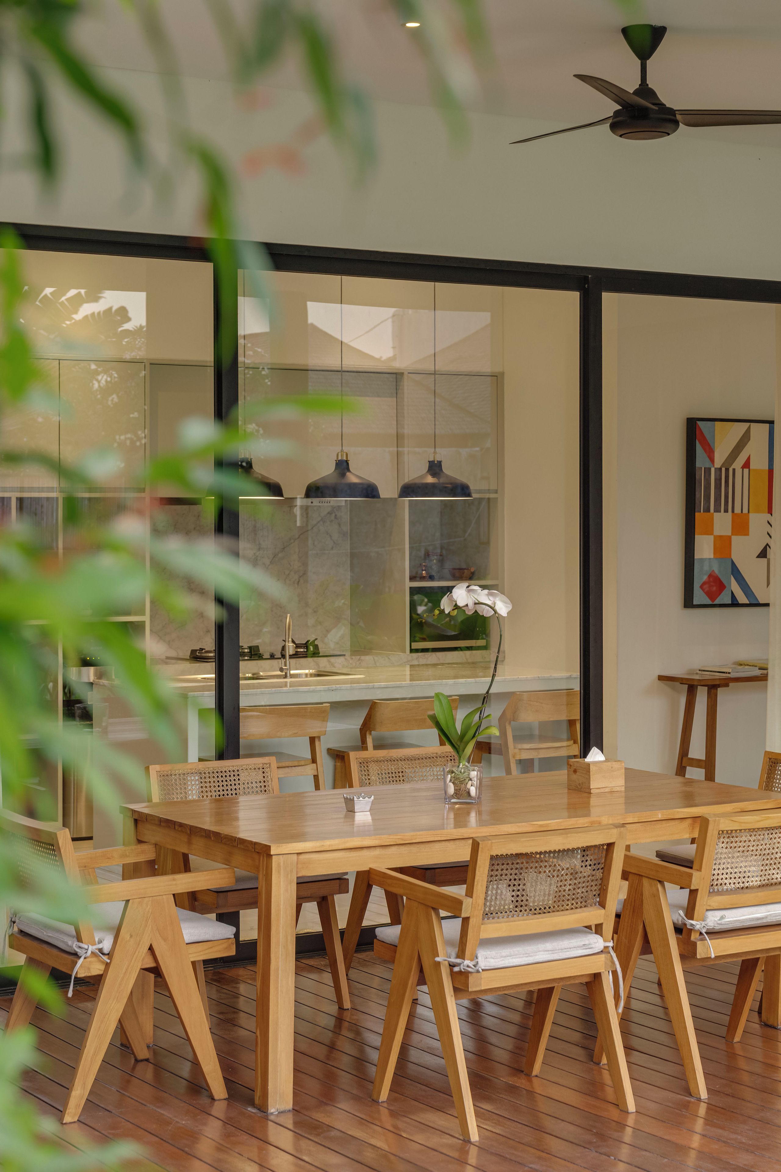 Comedor con mesa y sillas de madera, ventilador de techo y vista a través de un panel de vidrio.