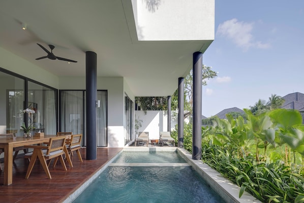 A modern outdoor pool area with a wooden deck, seating, and a view of the surrounding landscape. - VOX Residences Canggu (Tibubeneng)