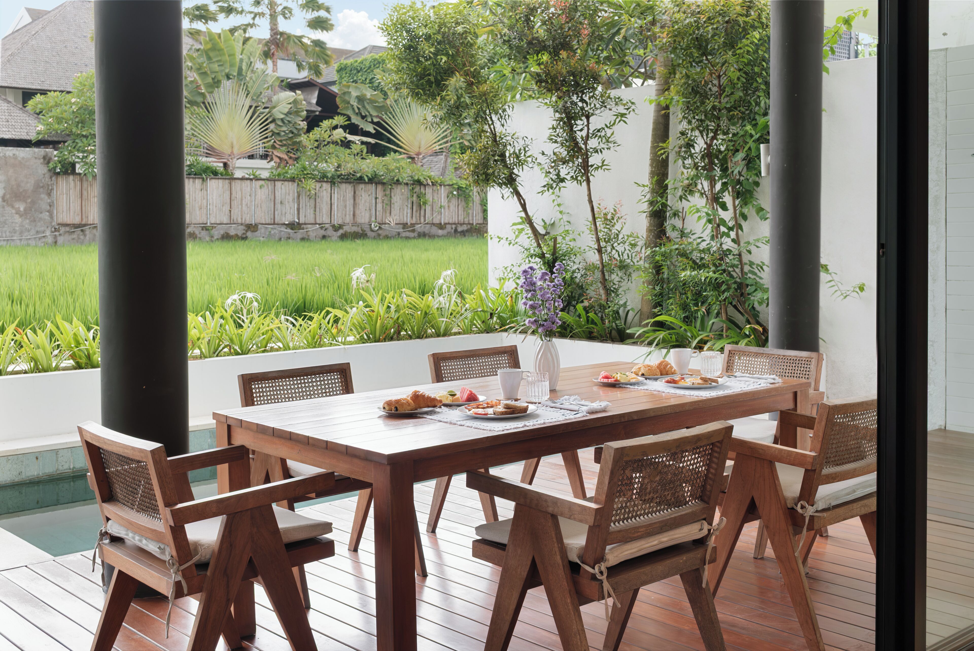 A wooden dining table with chairs, a vase with flowers, and a view of a garden and pool.