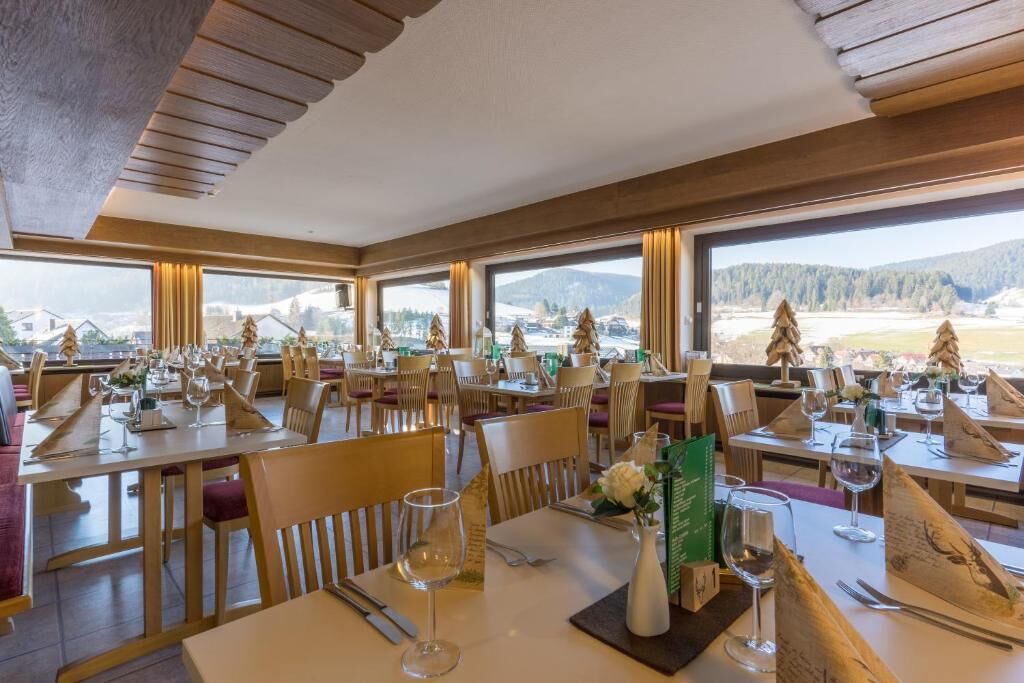 Ein Restaurant mit mehreren gedeckten Tischen, großen Fenstern mit Blick auf eine verschneite Landschaft und einer Holzbalkendecke.