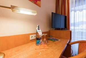 Habitación de hotel con escritorio de madera, un dispensador de gel antibacterial, dos copas de vino, un televisor y una ventana con cortinas.