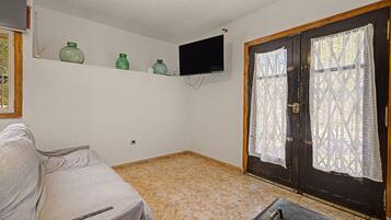 A room with a sofa, a television mounted on the wall, a door with lace curtains, and a tiled floor.