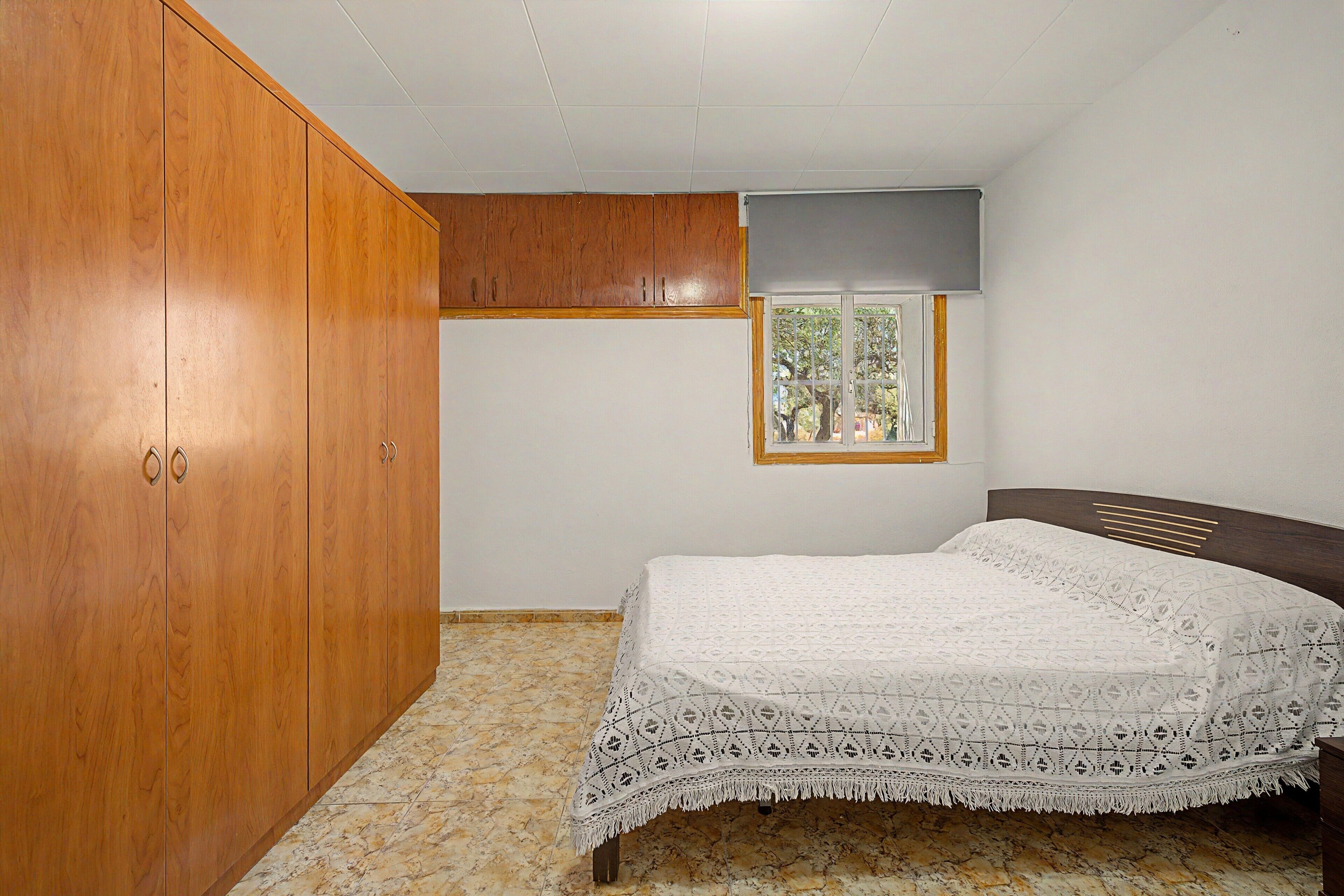 A bedroom with a wooden wardrobe, a bed with a patterned bedspread, a window with a view of trees, and a ceiling with recessed lighting.