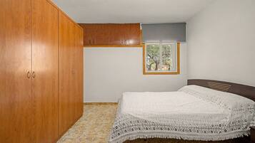 A bedroom with a wooden wardrobe, a bed with a patterned bedspread, a window with a view of trees, and a ceiling with recessed lighting.