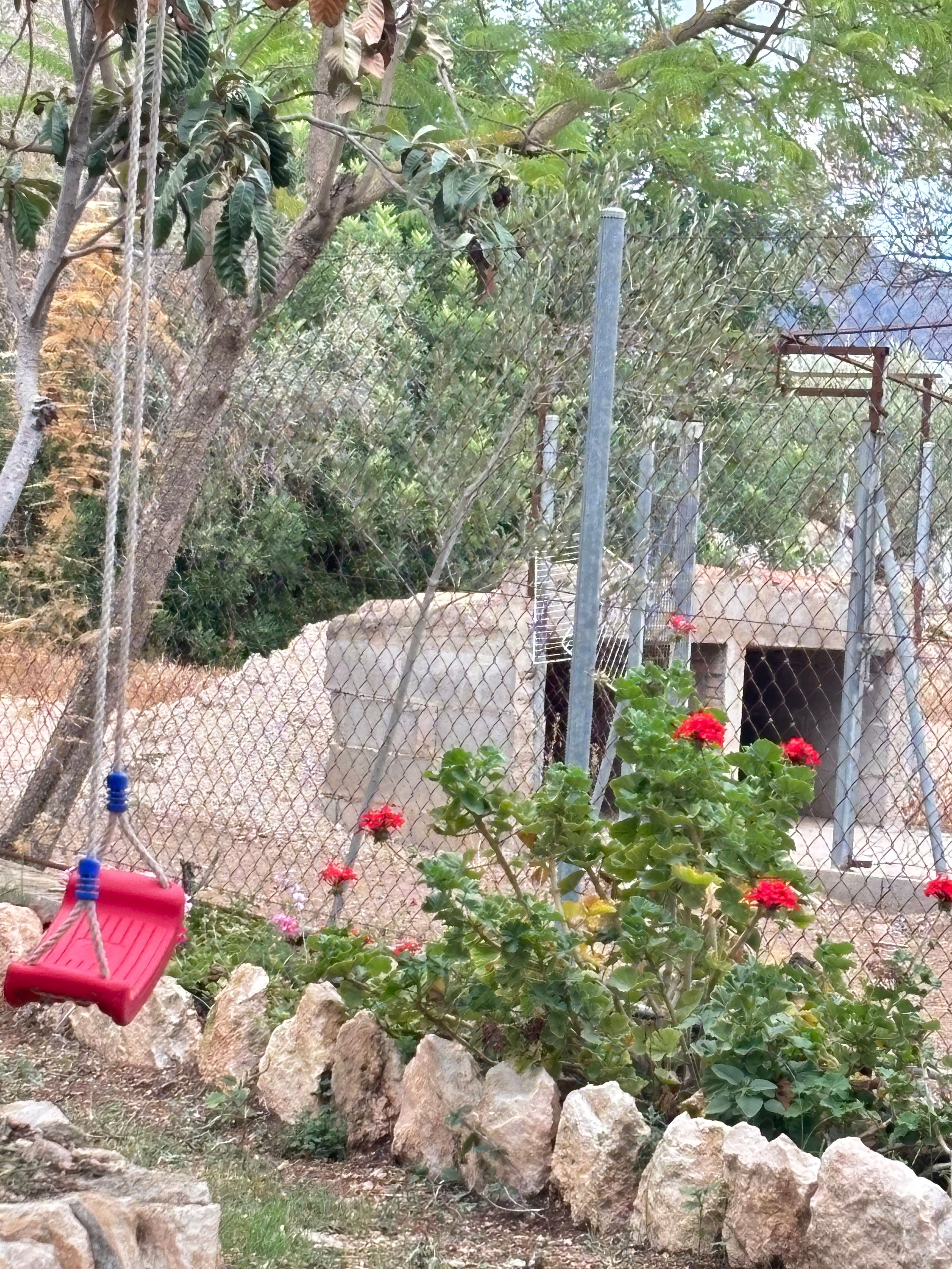 A fenced outdoor area with a swing, rocks, and a building in the background.